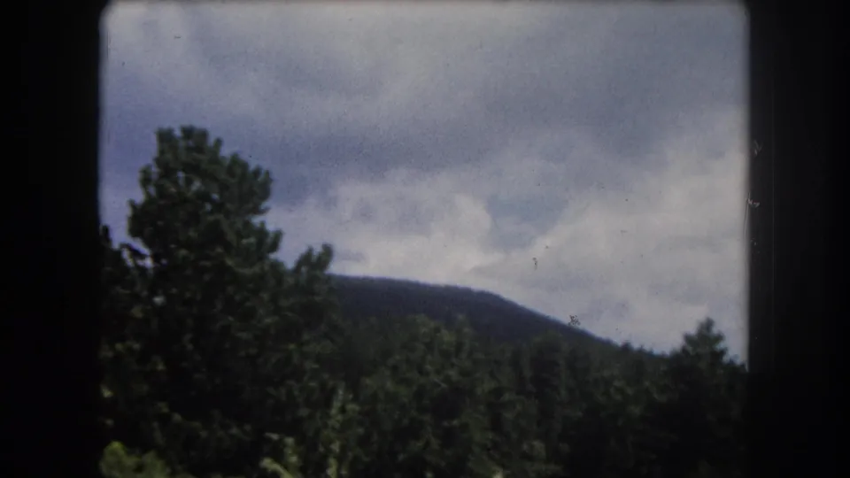 8mm home movie frame from 1962 showing conifer forest and rolling mountain ridge under cumulus clouds in Summit County, Colorado.