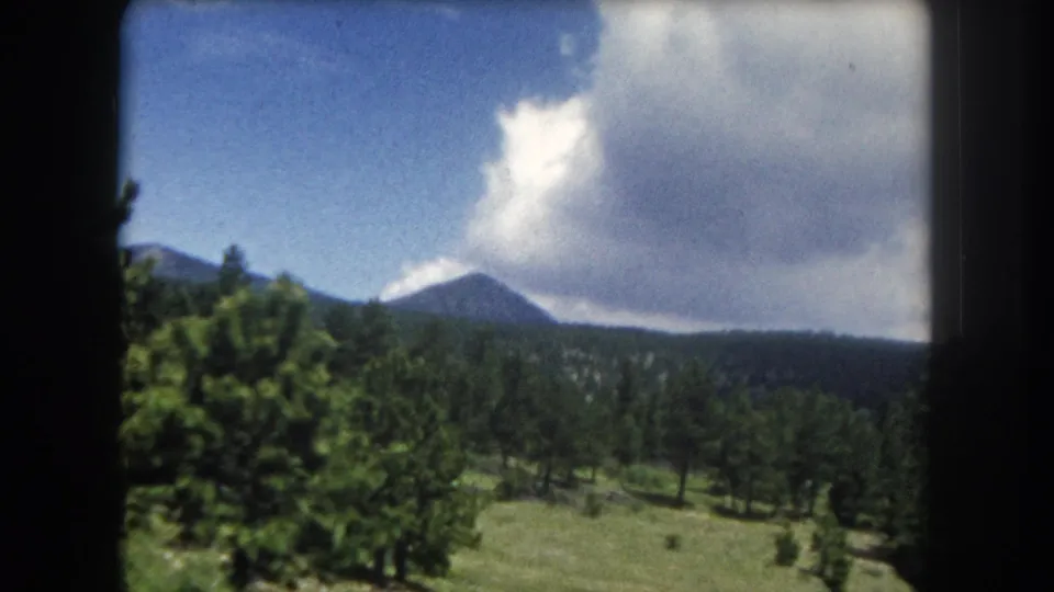 8mm home movie frame from 1962 showing a prominent mountain peak, pine forest, and open meadow under summer cumulus clouds in Summit County, Colorado.