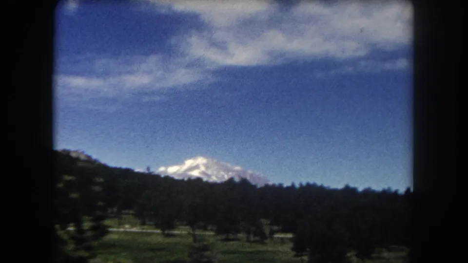 8mm home movie, c.1962, Summit County Colorado — snow-capped mountain above forested ridge, open meadow in foreground, deep blue sky with wispy clouds.
