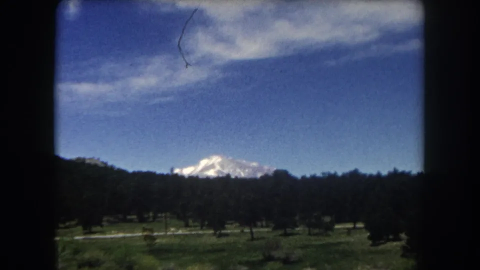 8mm home movie, c.1962, Summit County Colorado — snow-capped peak above evergreen ridge and meadow; film emulsion scratch artifact in upper right corner.