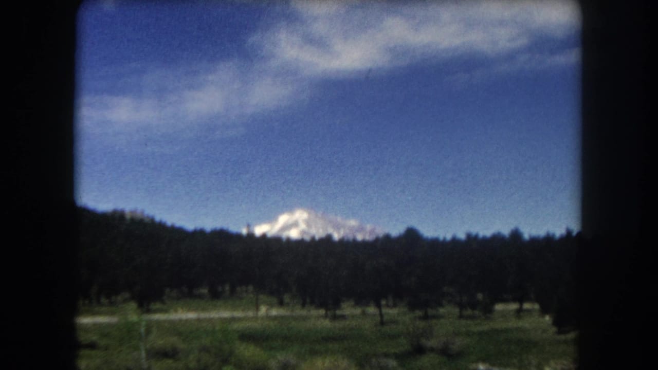 8mm home movie frame, c.1962 Summit County Colorado — snow-capped peak beyond conifer forest and open meadow, viewed from a moving car window.