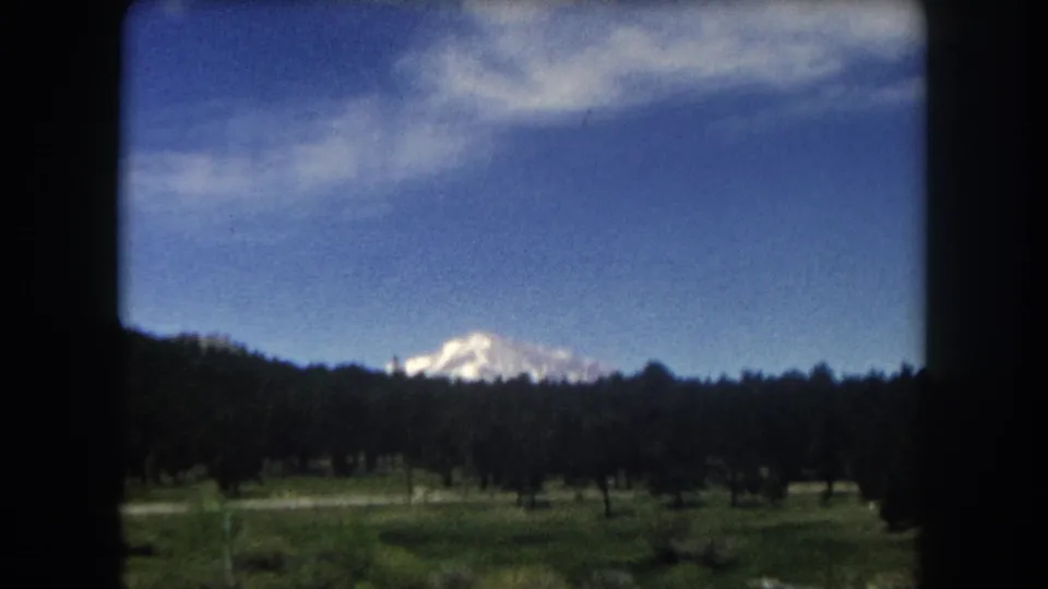 8mm home movie frame, c.1962 Summit County Colorado — snow-capped peak beyond conifer forest and open meadow, viewed from a moving car window.