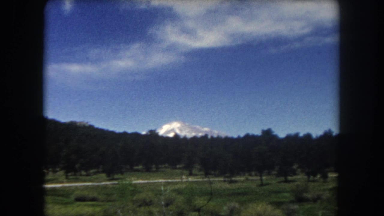 8mm home movie frame, c.1962 Summit County Colorado — mountain peak drifted rightward as car continues along road, conifers in midground.