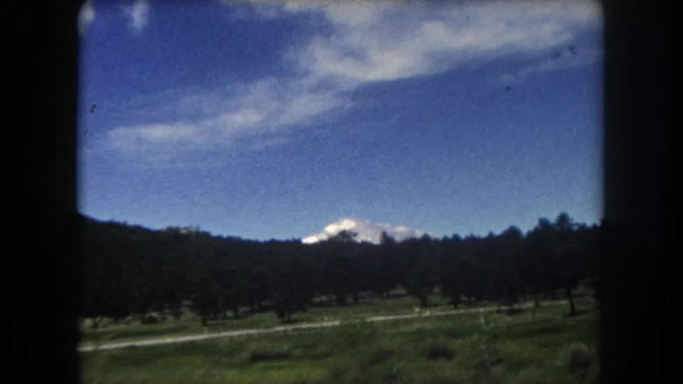 8mm home movie frame, c.1962 Summit County Colorado — camera angle rises, sky fills upper half, snow-capped peak partly obscured by conifer treeline.