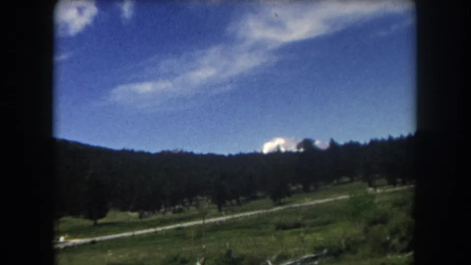 8mm home movie frame, c.1962 Summit County Colorado — snow-capped peak at far right as vehicle passes; treeline and meadow in mid-ground.