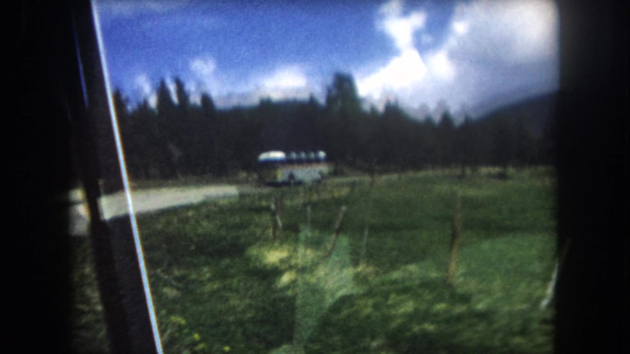8mm home movie from a moving car showing a mid-century coach bus rounding a curve on a Colorado mountain highway, pine forest and cumulus clouds visible, c. 1962.