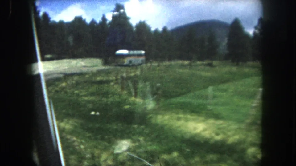 8mm home movie shot from a moving car of a mid-century bus traveling through a green Colorado mountain valley with pine forest and scattered clouds overhead, c. 1962.