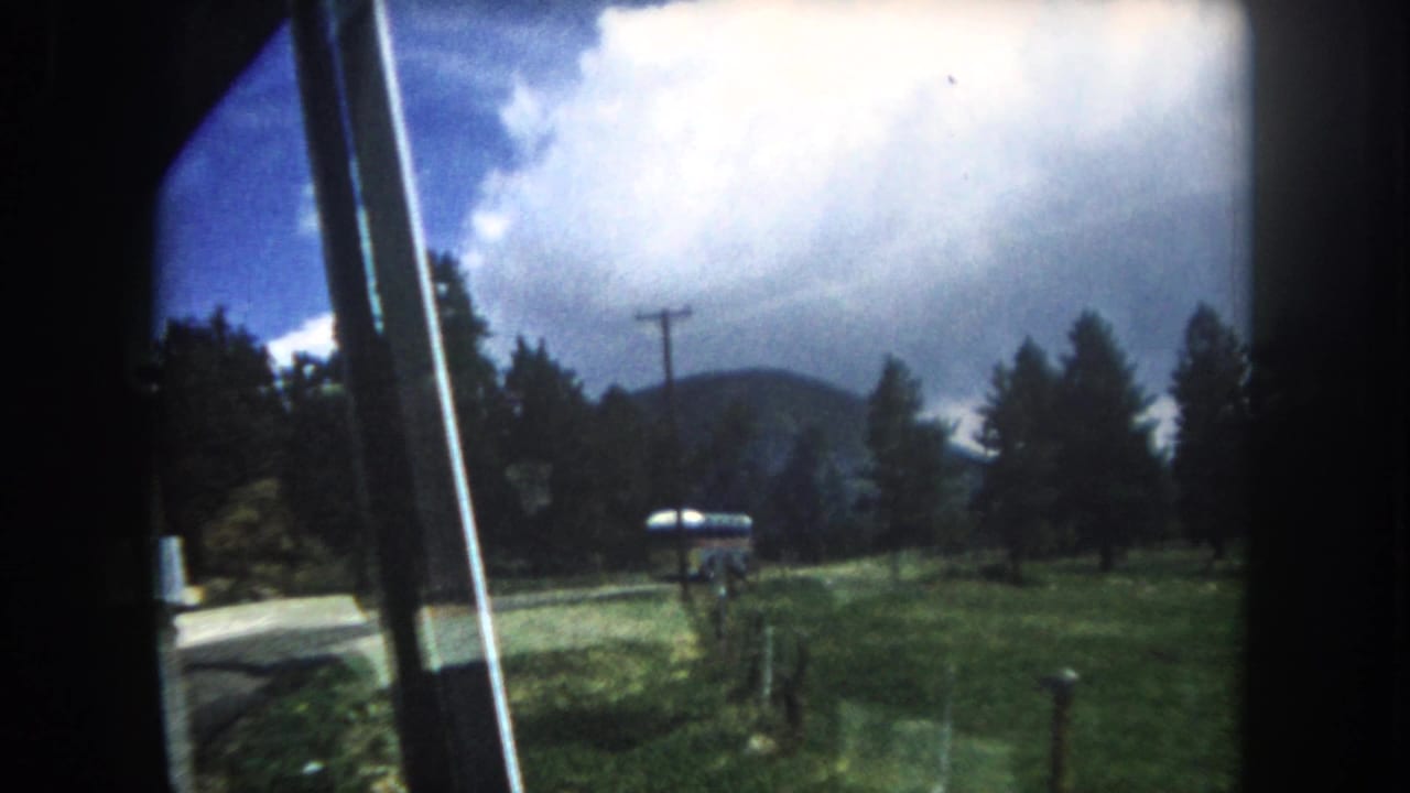 View from inside a car window showing a roadside bus, pine trees, a utility pole, and distant mountain hills under a blue sky.