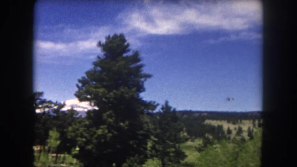 8mm film frame from inside a moving vehicle, c.1962 Summit County Colorado — prominent snow-capped peak rising behind conifer forest and green hills.
