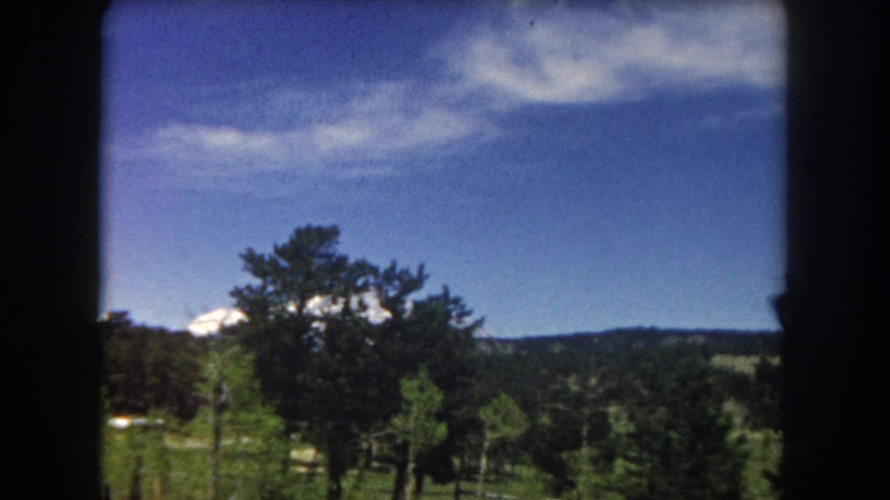 8mm home movie frame from 1962 showing a snow-capped Rocky Mountain peak rising above pine trees and a green valley in Summit County, Colorado, shot from a moving vehicle.