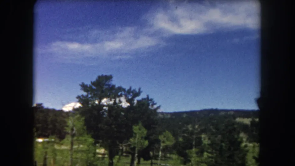 8mm home movie frame from 1962 showing a snow-capped Rocky Mountain peak rising above pine trees and a green valley in Summit County, Colorado, shot from a moving vehicle.