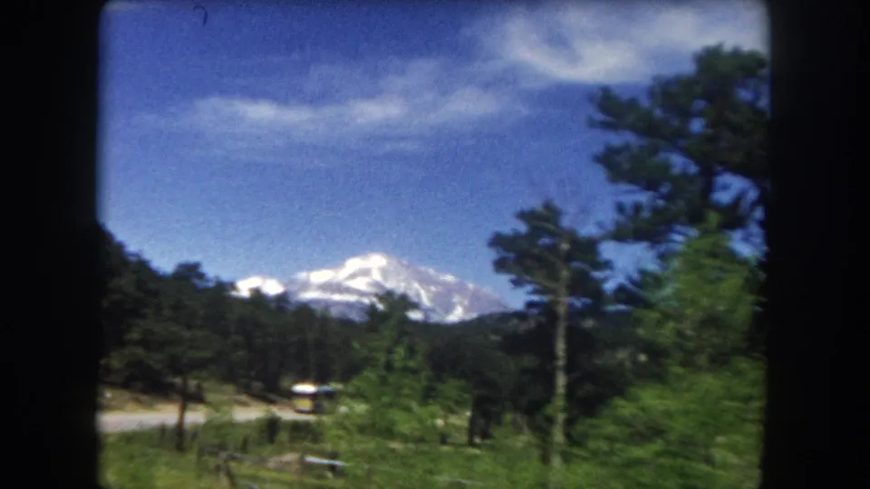 1962 8mm frame capturing a snow-capped Colorado peak framed by pine trees, with a valley road and distant vehicles visible below in Summit County.