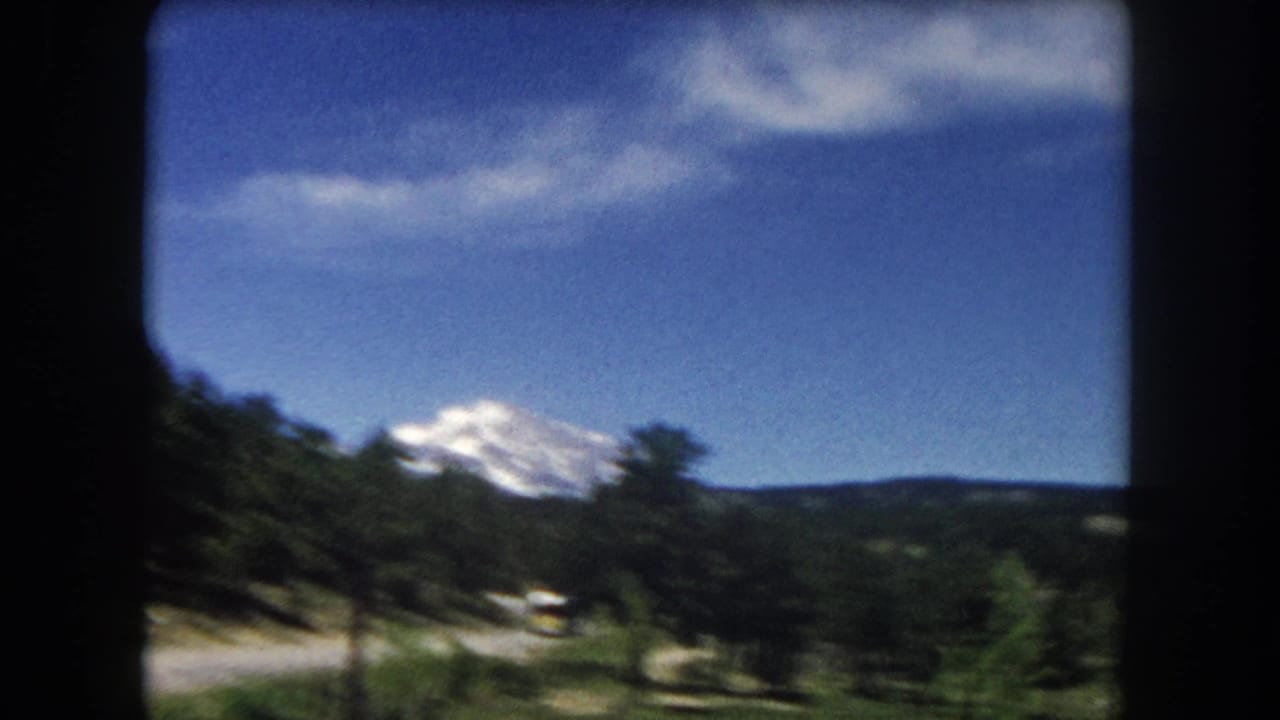 Forward vehicle-window view of paved mountain road curving through pine forest with snow-capped peak and yellow vehicle in distance, 8mm film.