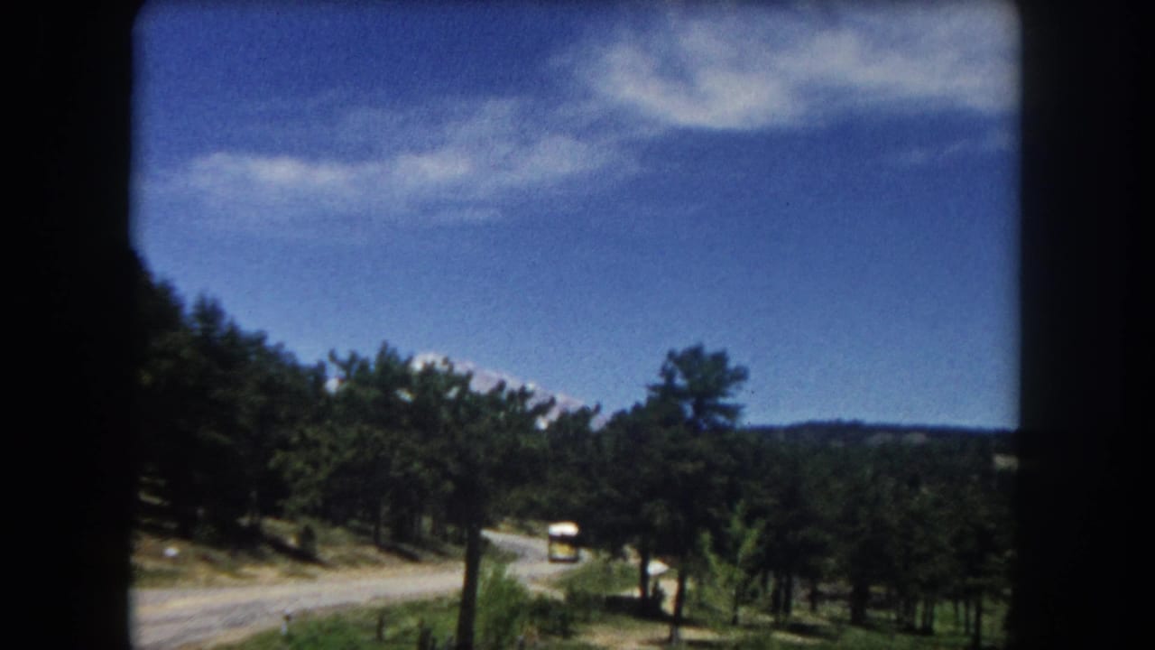 Vehicle-window view of winding mountain road with pine trees and yellow bus visible on right shoulder, 8mm film.