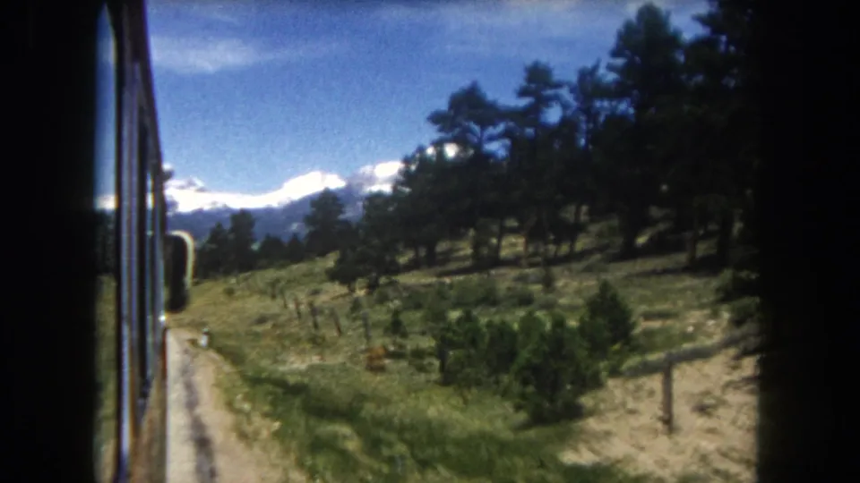 8mm home movie, circa 1962, Summit County Colorado: side-window perspective from moving vehicle — open grassland meadow with wire fence, scattered pines, and snow-capped mountain range.