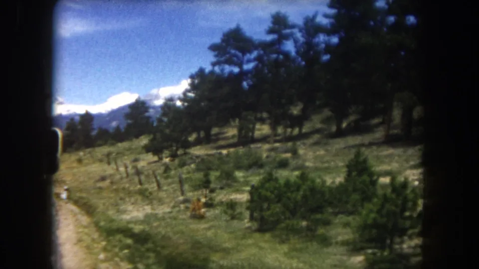 8mm home movie, circa 1962, Summit County Colorado: wide open meadow with sparse pines, wire fence, and snow-capped mountain range visible under a clear blue sky from a moving vehicle.
