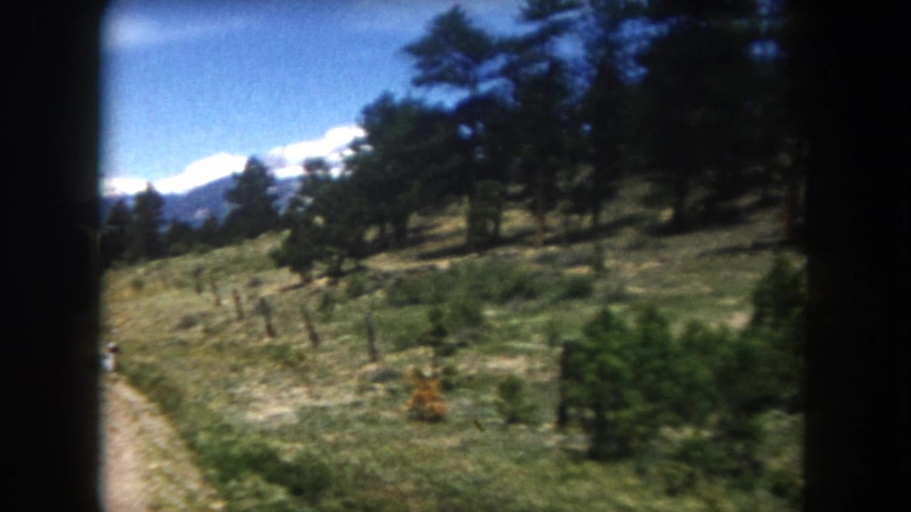 8mm view from a moving vehicle of a dirt road climbing through pines toward snow-capped Colorado Rockies peaks, circa 1962.