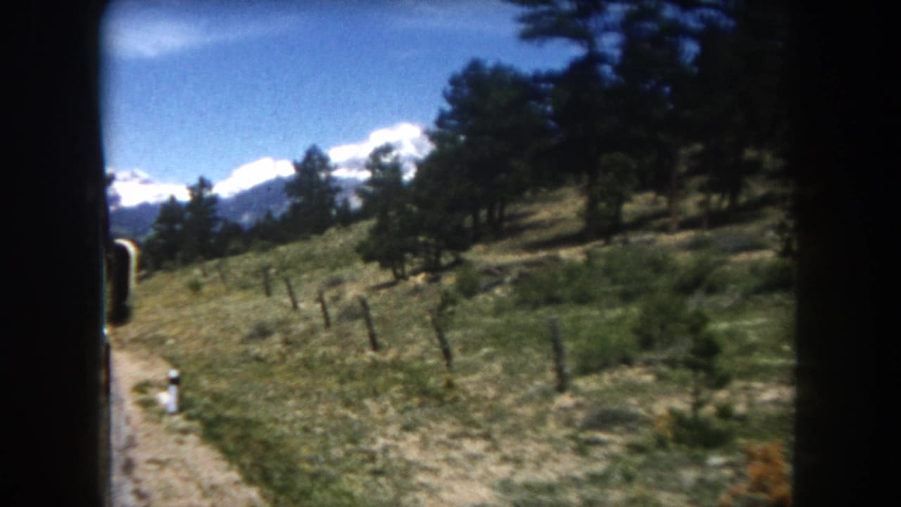 8mm side-window shot from a moving vehicle on a dirt mountain road in Summit County, Colorado, circa 1962, with snow-capped peaks and fence posts visible.