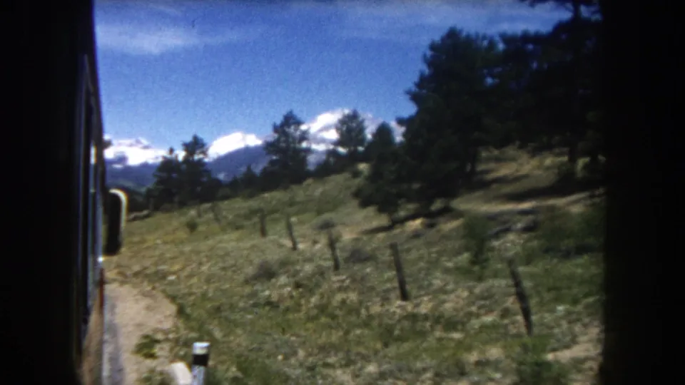 8mm footage through a vehicle window—possibly a school bus—showing a dirt road, post fence, and snow-capped Rocky Mountain peaks in Summit County, Colorado, circa 1962.