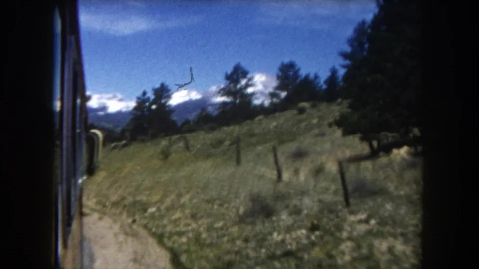Final 8mm frame from a moving vehicle in Summit County, Colorado, circa 1962, showing the road curving past a fence line with snow-capped Rockies in the background.