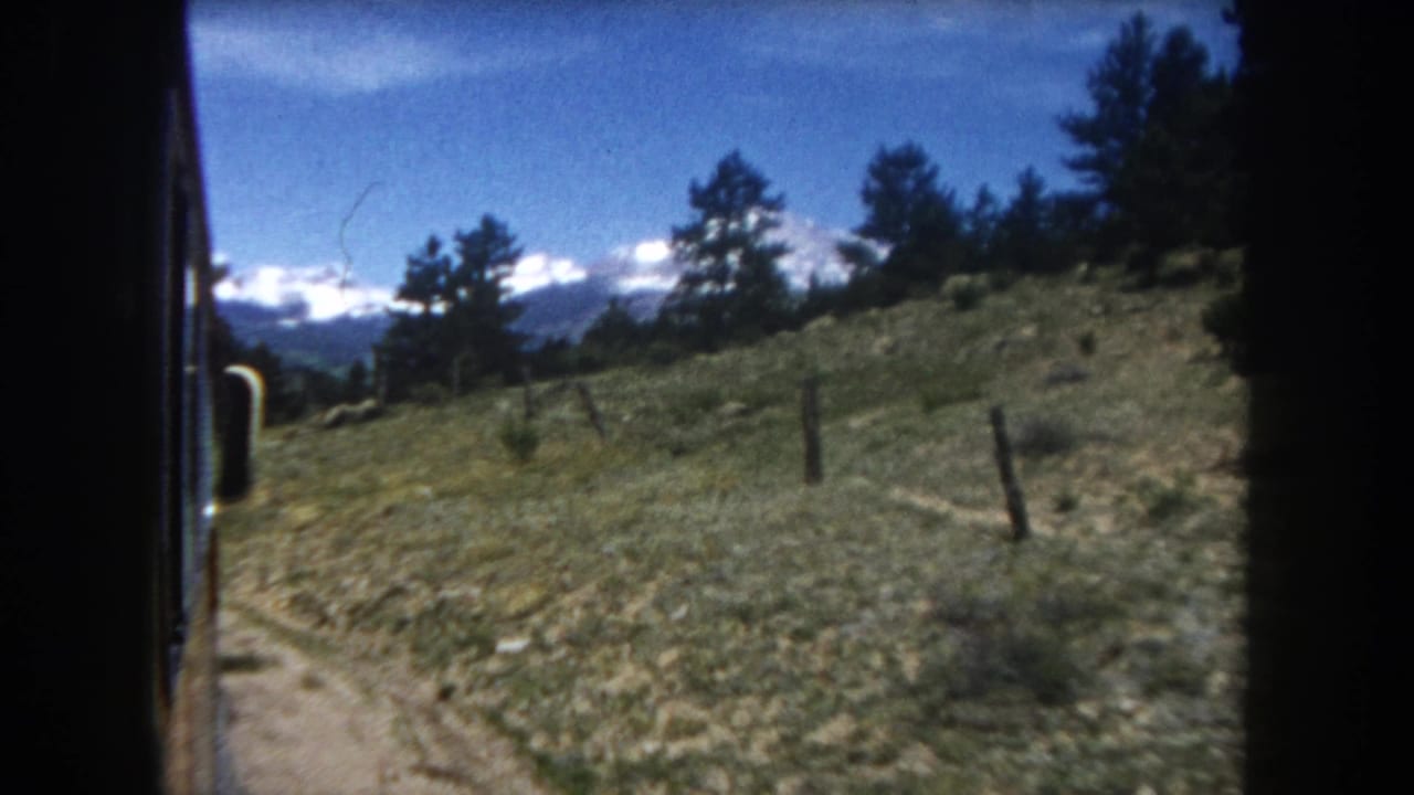 1962 8mm home movie: looking out a Colorado train window at a green hillside, fence post, pine trees, and distant snow-capped Rocky Mountains.