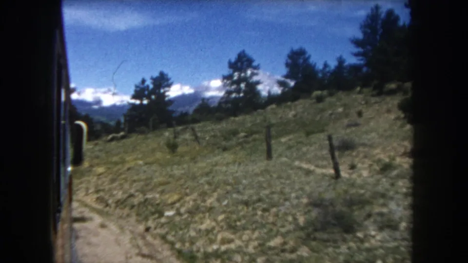 1962 8mm home movie: looking out a Colorado train window at a green hillside, fence post, pine trees, and distant snow-capped Rocky Mountains.