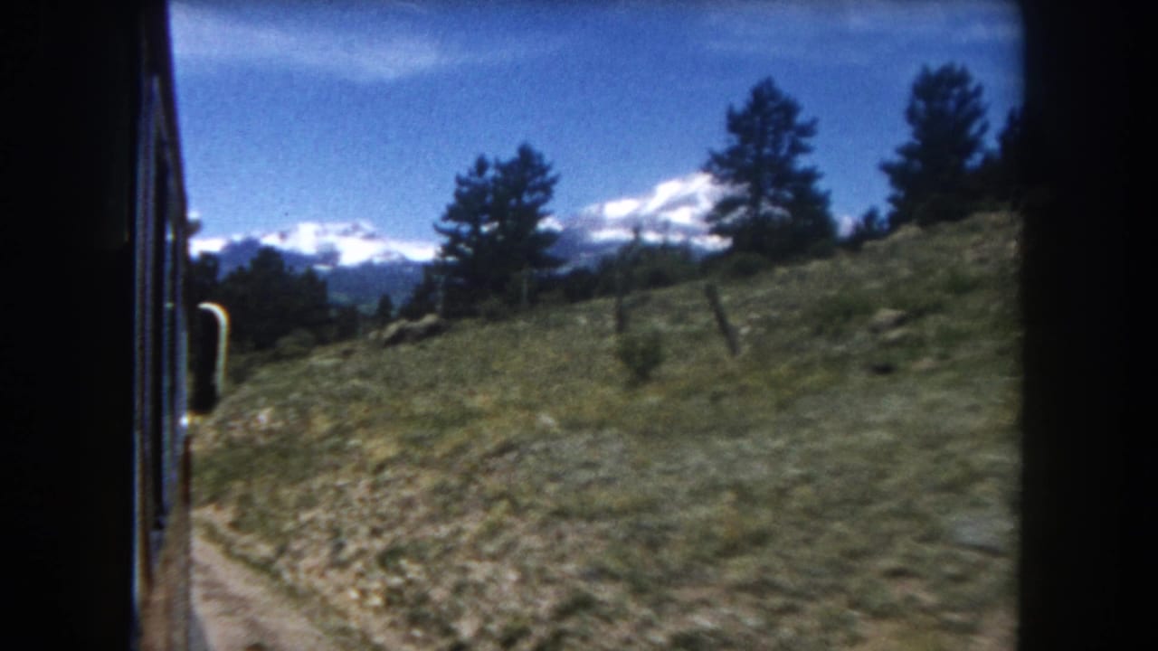 Ongoing 1962 Colorado train journey footage: hillside vegetation, fence post, and distant Rocky Mountain snow peaks framed by conifers.