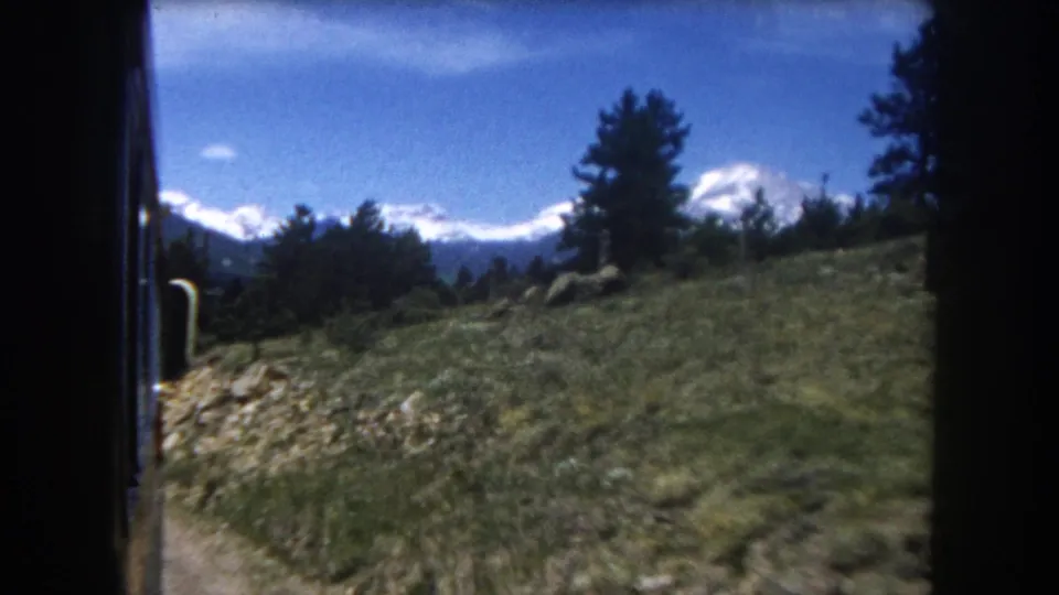 8mm Colorado mountain train footage, 1962: conifers, rocky terrain, and snow-covered peaks under a partly cloudy sky.