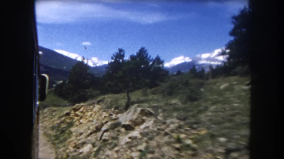 Final frame of 1962 Colorado train journey: rocky outcrop, conifers, and a snow-capped Rocky Mountain peak under blue sky.