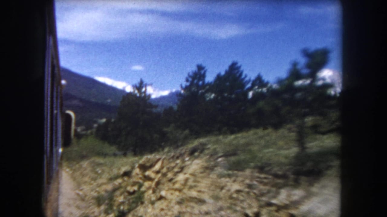 8mm home movie frame, c.1962, Summit County Colorado: dirt road lined with rocks and pine trees, snow-capped Rocky Mountain peaks visible in background, shot from car window.