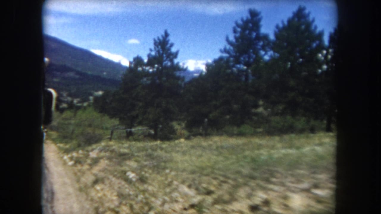 8mm frame, c.1962, Summit County Colorado: dirt road through green alpine meadow, dense pine trees at mid-frame, snow-capped mountain range on the horizon.