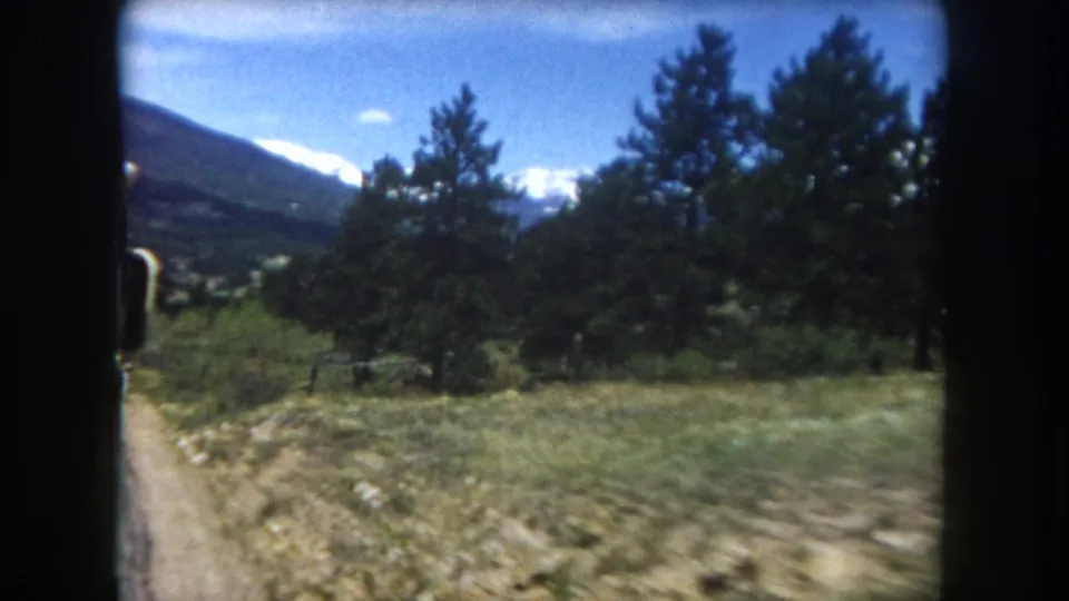 8mm frame, c.1962, Summit County Colorado: dirt road through green alpine meadow, dense pine trees at mid-frame, snow-capped mountain range on the horizon.