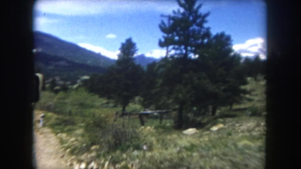 8mm frame, c.1962, Summit County Colorado: open alpine meadow shot from car window, scattered pines, snow-capped Rocky Mountain range beyond, wooden fence and distant white marker visible.