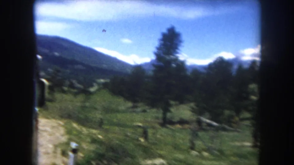 8mm frame, c.1962, Summit County Colorado: wide mountain valley from car window, pine trees flanking the scene, snow-covered Rocky Mountain peaks dominating the horizon with lens flare at right.