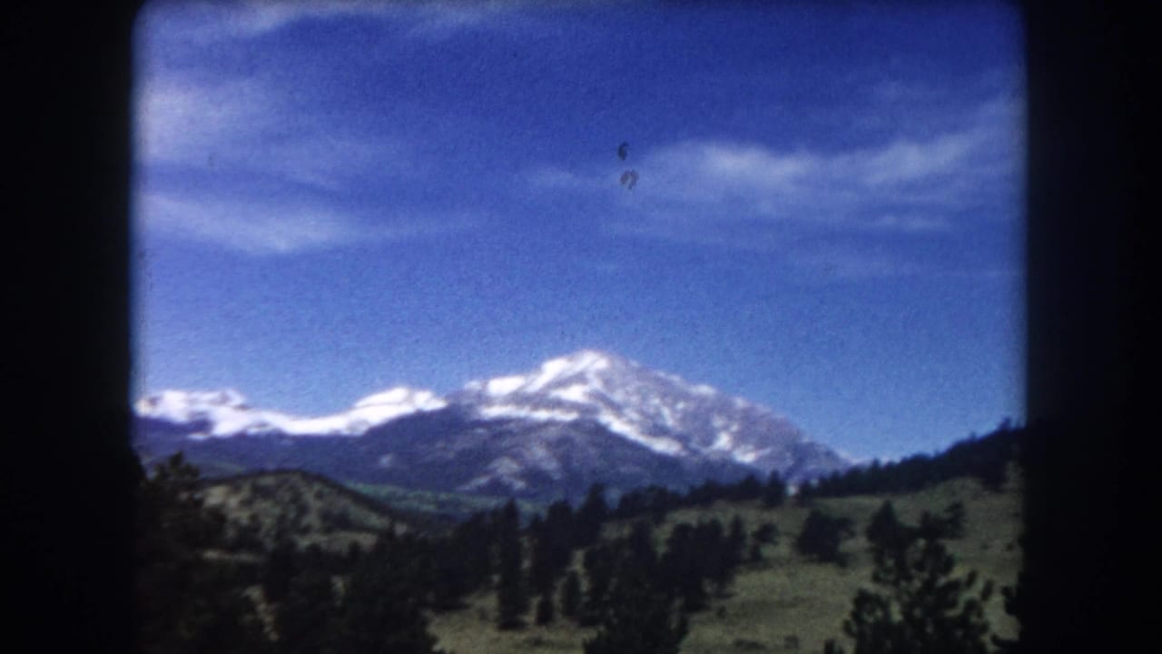 Snow-capped mountain peak over green foothills and pine trees, 8mm film borders visible.