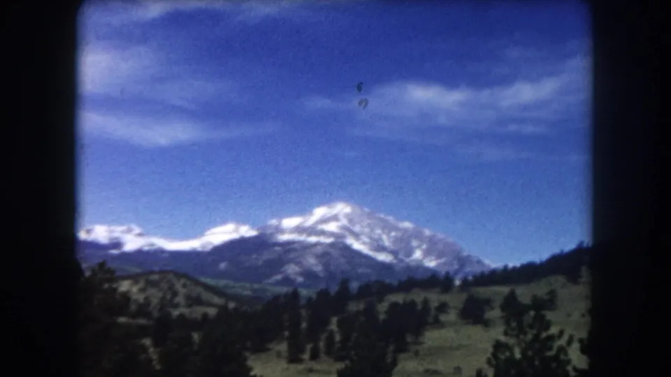 8mm film frame circa 1962, Summit County Colorado — snow-capped peak, green foothills, pine trees, blue sky with wispy clouds.