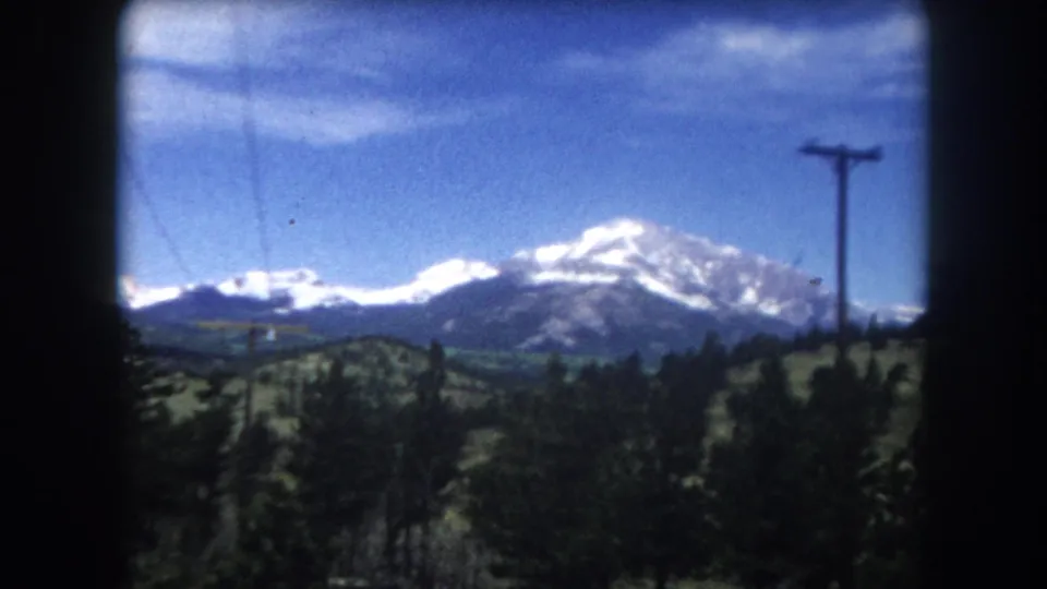 8mm film frame circa 1962, Summit County Colorado — snow-capped peak, green valley, foreground pines, and a utility pole visible in the mid-ground.