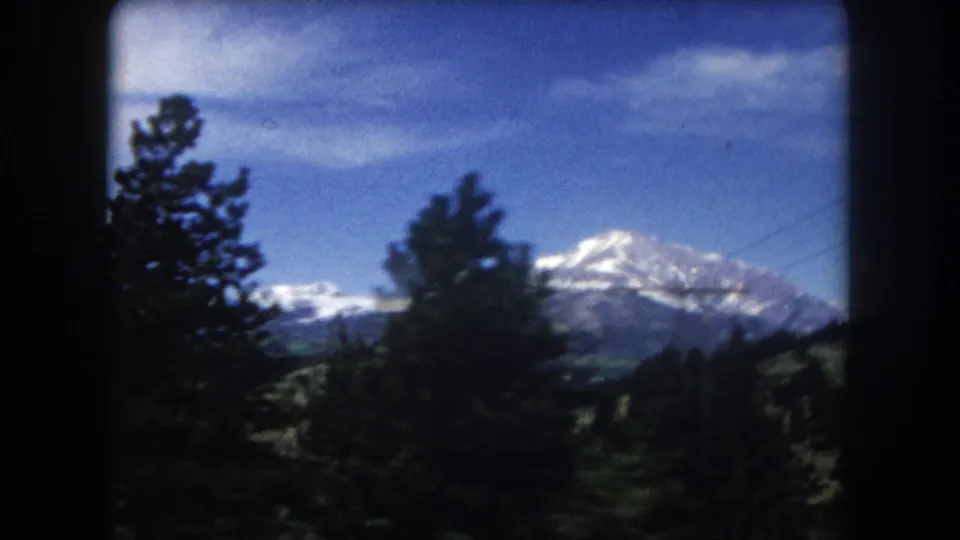 8mm film frame circa 1962, Summit County Colorado — dark foreground pine trees frame a snow-capped mountain peak visible through the branches.