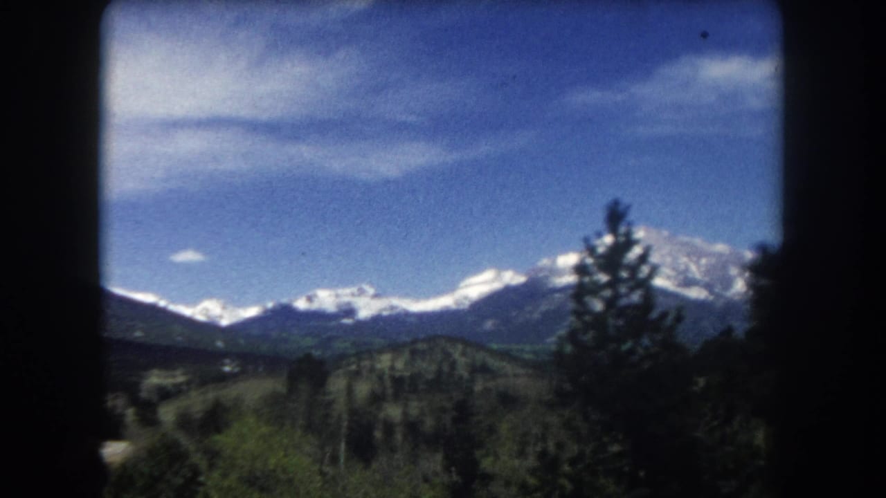 8mm home movie frame circa 1962 showing a snow-capped Colorado Rocky Mountain range under blue sky with wispy clouds and conifer trees at the frame edge.