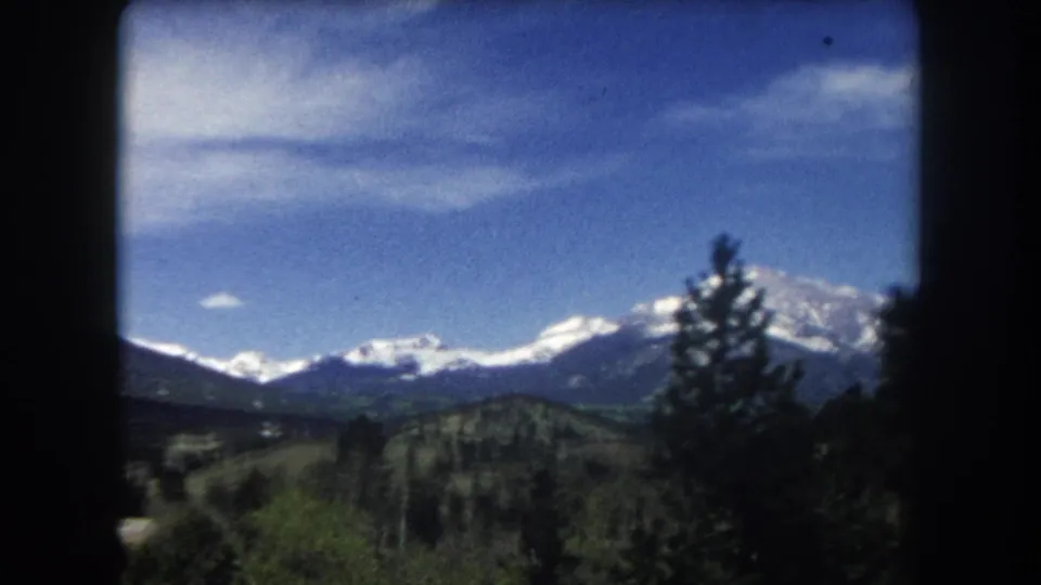 8mm home movie frame circa 1962 showing a snow-capped Colorado Rocky Mountain range under blue sky with wispy clouds and conifer trees at the frame edge.