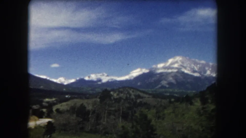 8mm home movie frame from 1962 showing a winding road through an open valley in Summit County, Colorado, with a large snow-capped peak rising behind rolling hills.