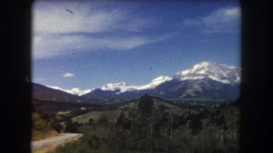 8mm home movie frame from 1962 showing a road through Summit County, Colorado with snow-capped peaks and open terrain under a blue sky.