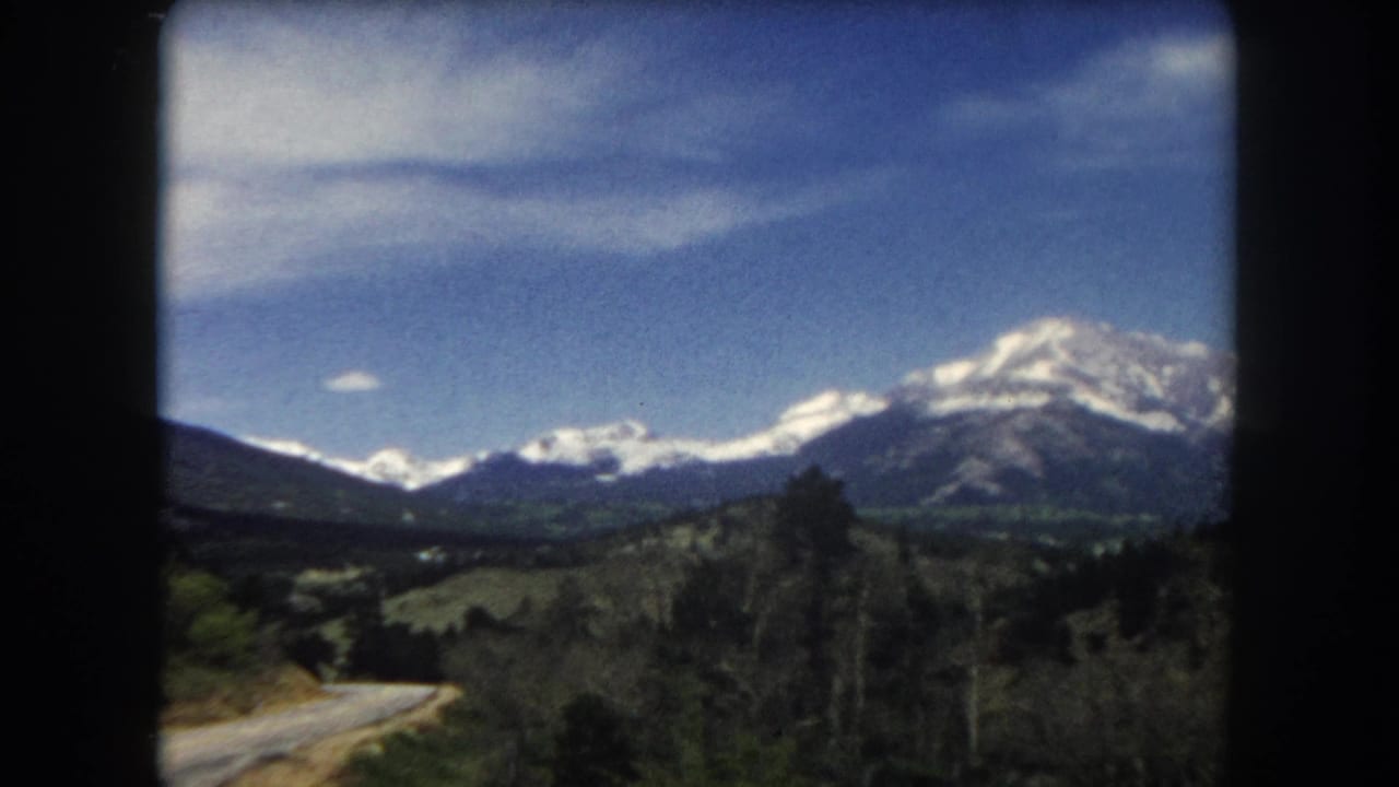 Snow-capped mountains across rolling green hills with a road in the lower foreground, viewed through 8mm film grain.