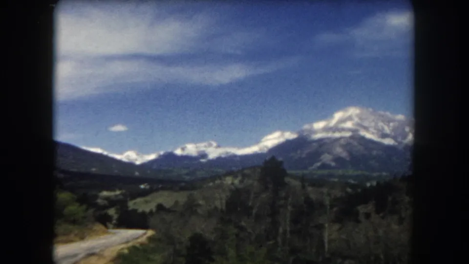 8mm home movie frame from c. 1962 showing a snow-capped mountain range over rolling green hills and road in Summit County, Colorado.