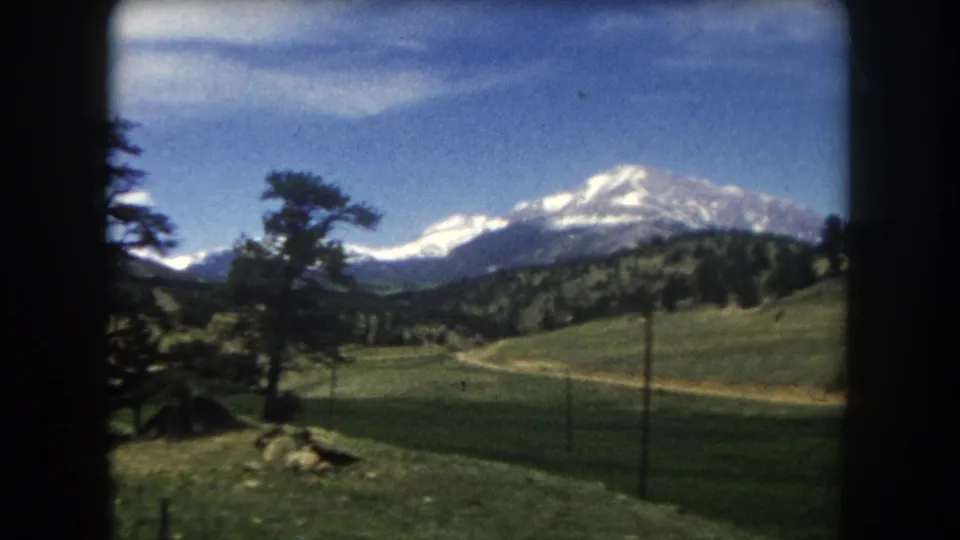8mm home movie frame c. 1962 showing snow peaks over green pasture with pine trees and a roadside utility pole, likely Summit County, Colorado.