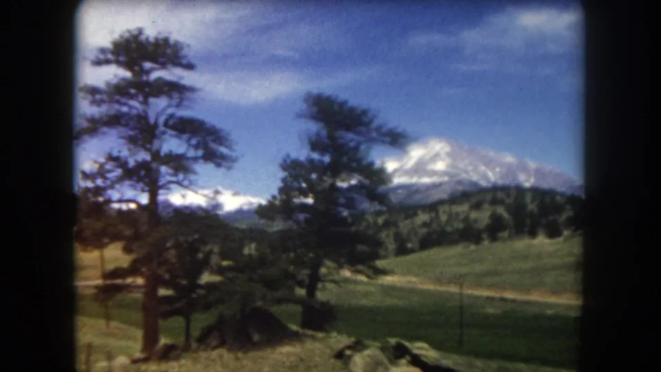 8mm home movie frame c. 1962 with large pine trees framing a snow-capped mountain and open meadow, likely Summit County, Colorado.