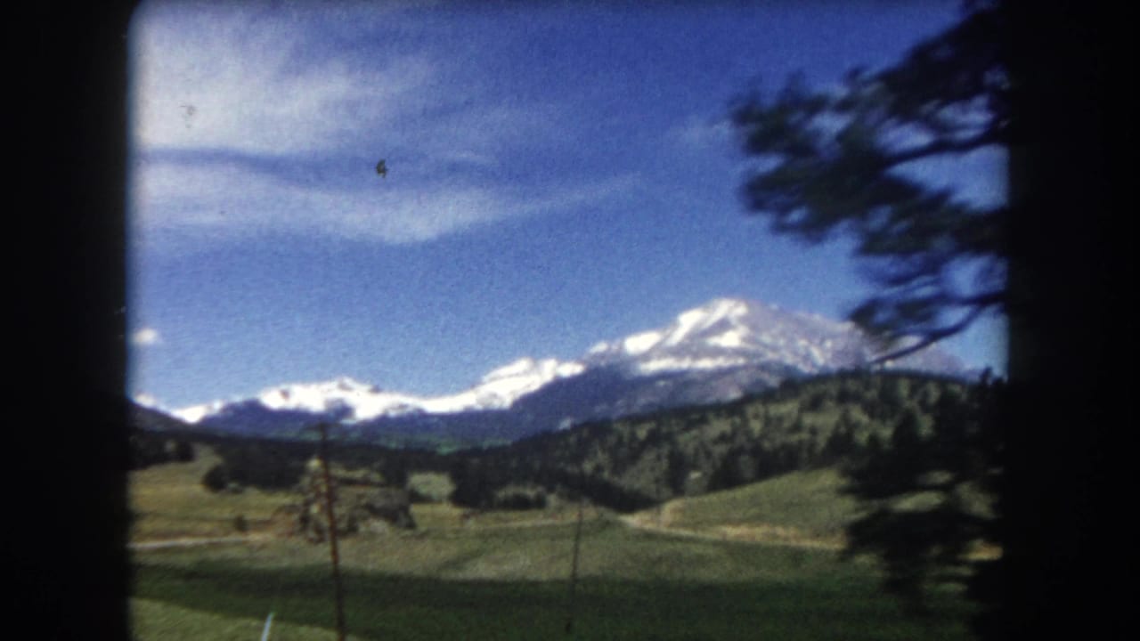 8mm home movie frame from 1962 showing snow-capped mountains in Summit County, Colorado, shot from a moving vehicle with power lines and evergreen trees visible.