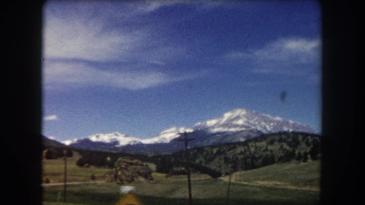 Open view of snow-capped peaks across green hills with wooden power line poles and rocky outcroppings in the mid-ground under a blue sky.