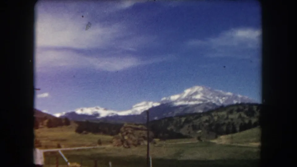 8mm home movie frame from c. 1962 showing a snow-capped Colorado peak under a violet-shifted sky, with power lines, rolling hills, and roadside infrastructure.