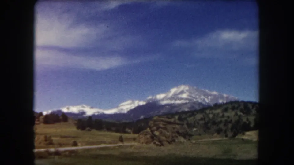 Mid-pan 8mm frame from Summit County, Colorado, 1962; snow-capped mountain range over green hills and scattered conifers under blue sky.